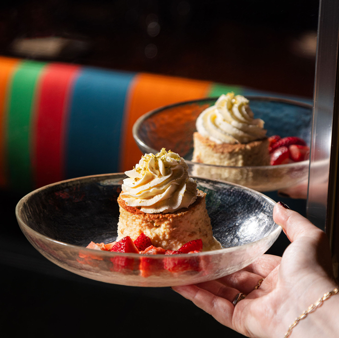 A hand holds a glass plate with a small dessert topped with whipped cream and garnished with strawberries.