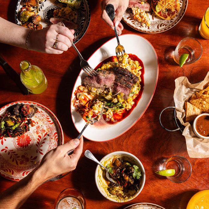 Top-down view of wooden table with vibrant dishes, including steak, sides, and drinks, in a colorful, ornate plate setting.