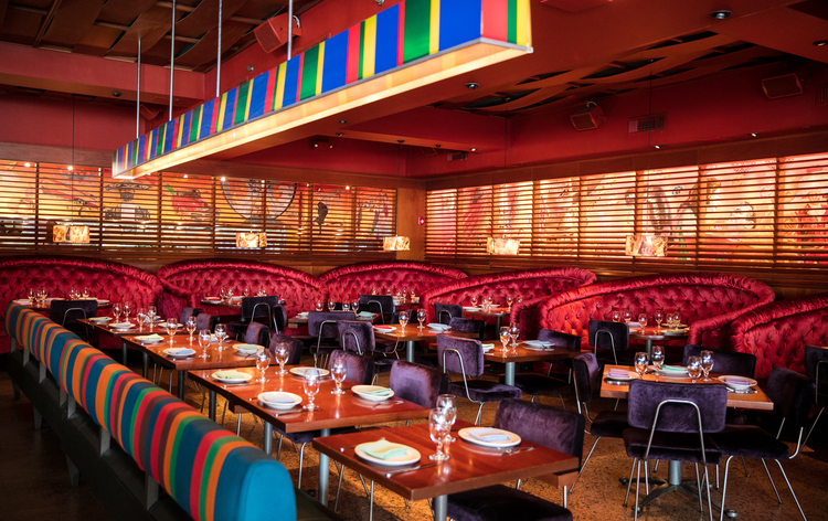 A cozy restaurant interior with red booth seats, colorful striped back cushions, wooden tables, and vibrant wall art through louvered windows.