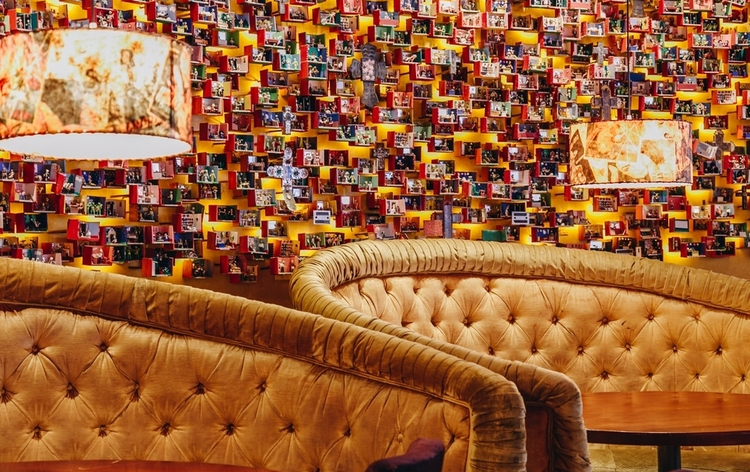 A cozy restaurant interior with red booth seats, colorful striped back cushions, wooden tables, and vibrant wall art through louvered windows.